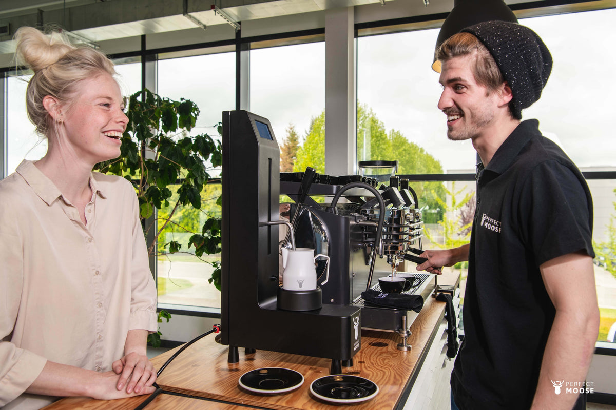 Barista serving a customer beside a black Perfect Moose steaming a white Smart Jug